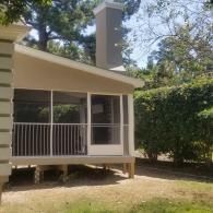 A tan screened-in porch with a white door and metal railing sits on a wooden deck next to a tall, grey chimney.