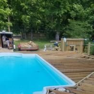 An outdoor swimming pool with a partially constructed wooden deck and building materials in a grassy, wooded yard.