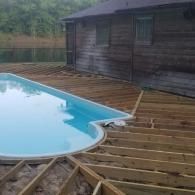 A wooden deck frame under construction surrounding a kidney-shaped, light blue swimming pool near a wooden house.