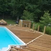 Wooden deck framing being built around a backyard swimming pool surrounded by trees.