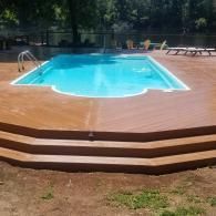 An outdoor swimming pool set into a light brown wood deck with three wide steps, surrounded by trees and a water view.