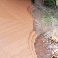 A high-angle view of a wooden deck with curved stairs leading down to a stone patio with outdoor wooden chairs.