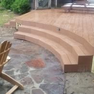 Curved, light brown wooden deck stairs leading up to a platform, set against a stone patio and a wooden chair.