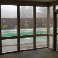 A view through a screened-in porch looking out at a swimming pool in a backyard with a wooden fence and foggy sky.