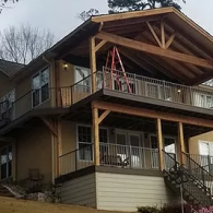 A two-story house with a wooden deck and balcony under construction, featuring an exposed beam roof and a metal ladder.