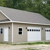 A tan vinyl-sided detached garage with white trim, a gray roof, two white garage doors, a side door, and a small window.