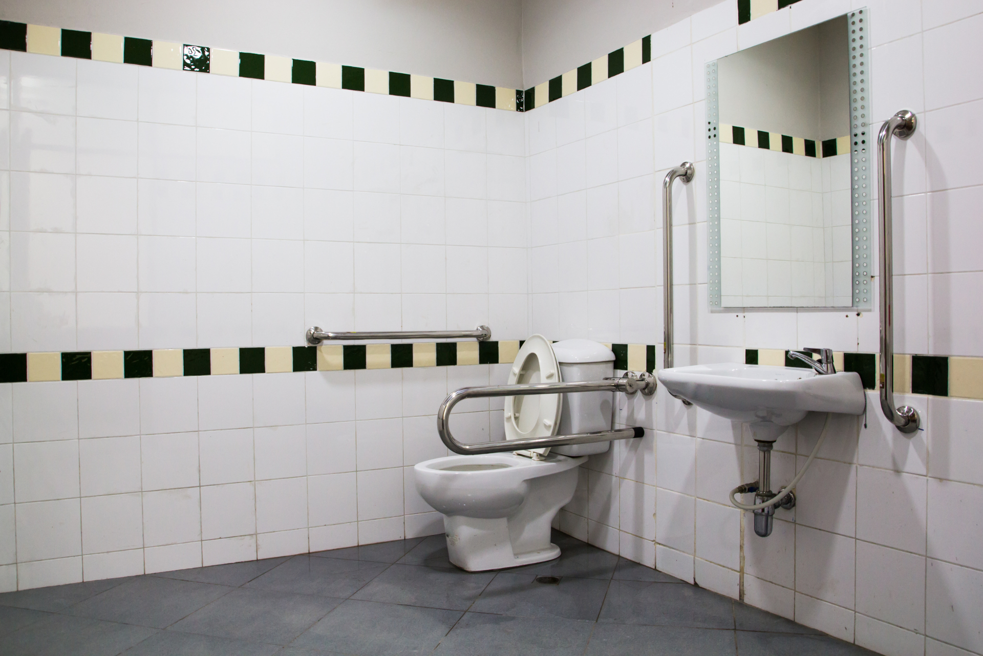 Toilettes publiques accessibles avec barres d'appui, toilettes, lavabo et miroir. Murs carrelés blancs avec bordures vertes et noires.