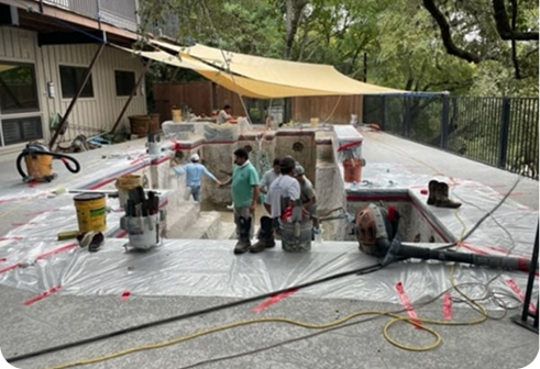 Construction workers in a pool renovation area with a tan sun shade, buckets, tools, and plastic sheeting on the ground.