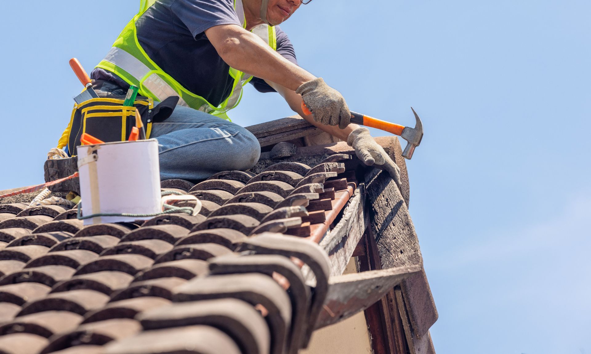 A man is kneeling on top of a tiled roof holding a hammer.