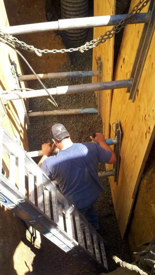 Worker in a hard hat standing on a ladder in a narrow trench beside yellow equipment, reaching into a wall panel.