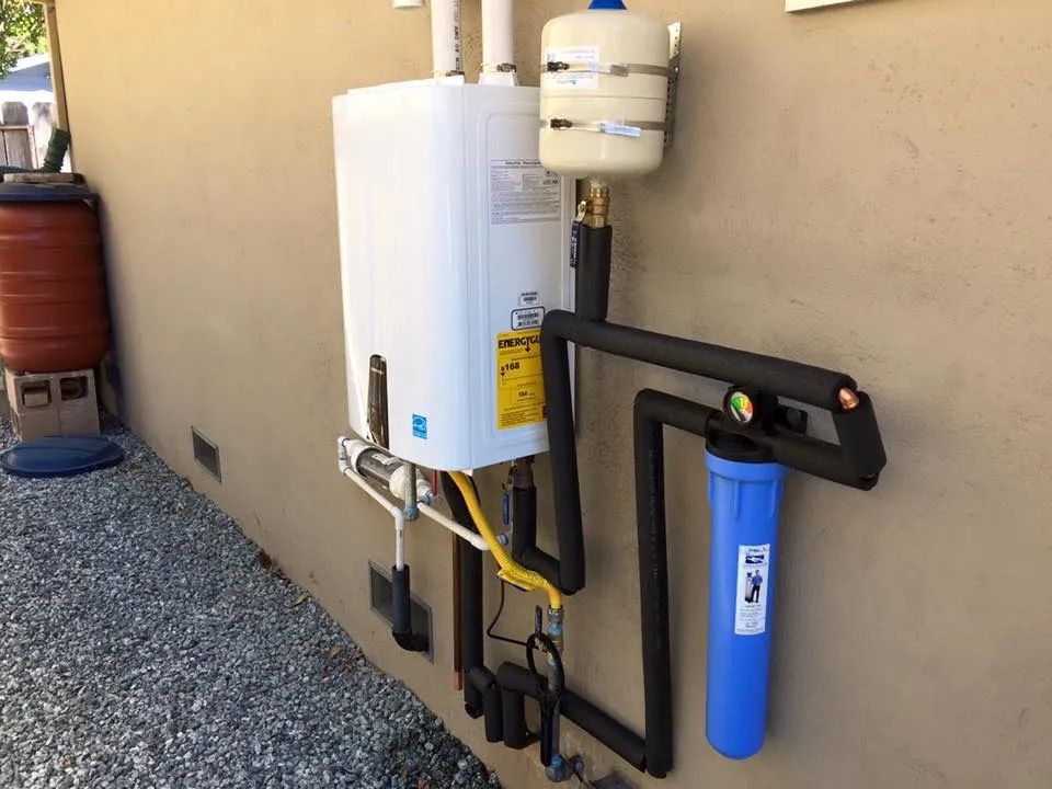 Wall-mounted water heater with blue filter and exposed pipes beside a gravel walkway.
