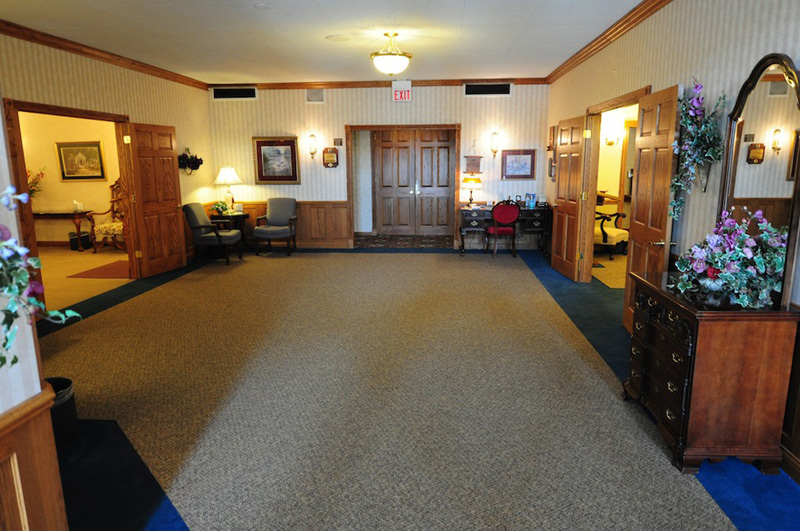Empty funeral home reception area with tan carpet, wood paneling, and doors.