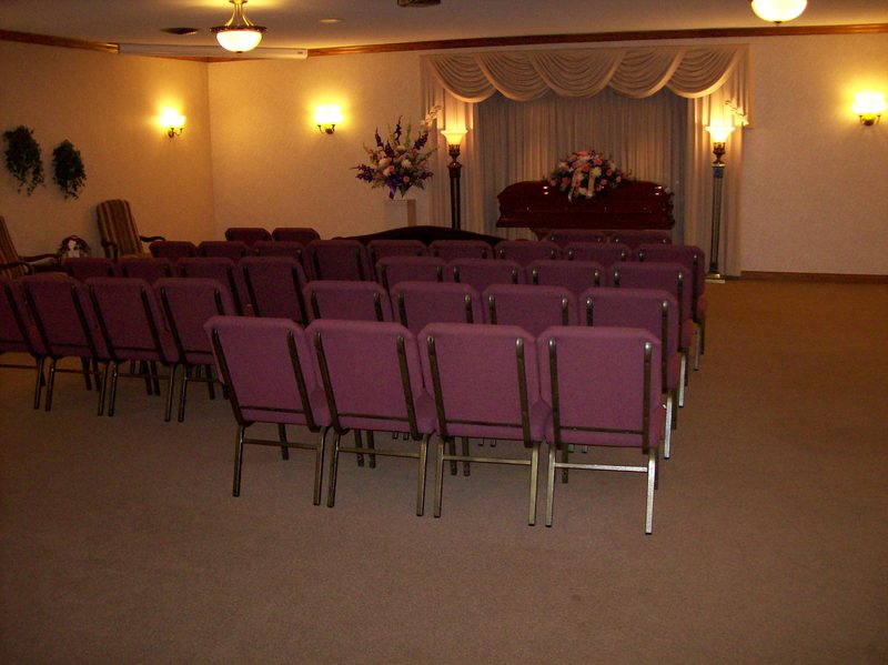 Interior of a funeral home with rows of purple chairs facing a casket. Flowers and lighting create a somber atmosphere.