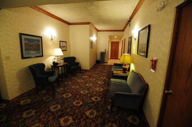 Hallway with patterned carpet, seating, artwork, and a brown door.
