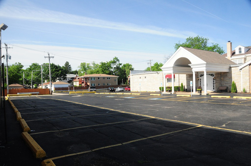 Empty asphalt parking lot next to a light-colored building with a covered entrance; blue sky.