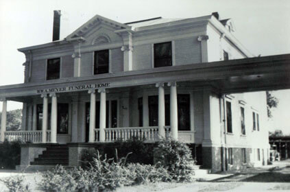 Exterior view of a two-story funeral home with columns and a porch, in black and white.