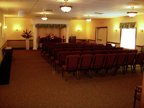 Funeral home interior with rows of chairs facing a floral arrangement.
