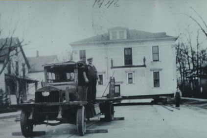 A large house being moved on a flatbed truck; a man stands on the truck.