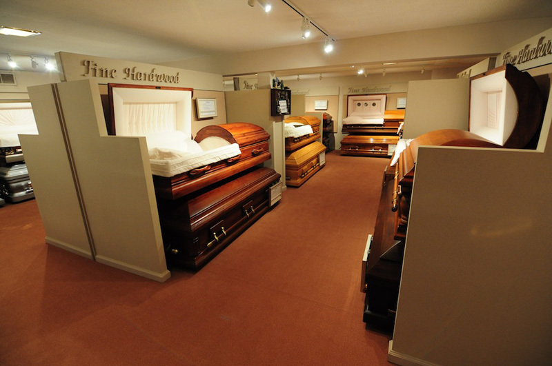 Casket showroom with various caskets on display, separated by white partitions. Reddish-brown floor, overhead lighting.