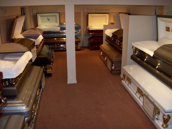 Room with several caskets in a funeral home. Various colors and materials, stacked and displayed.