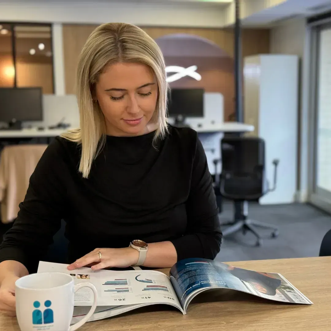 Woman in black top looking at a brochure at a desk, mug on the left.
