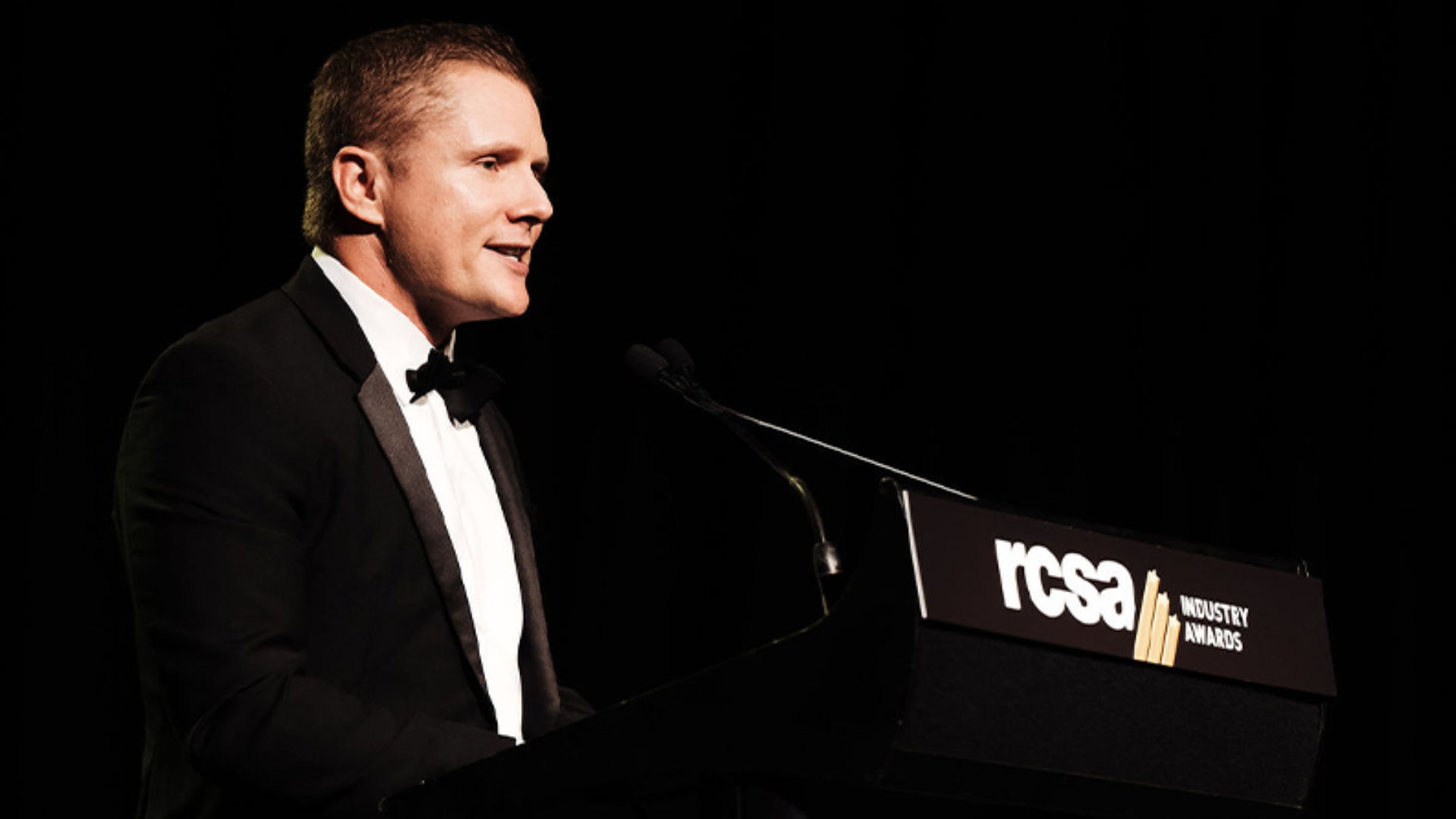 Man in tuxedo speaking at podium with 