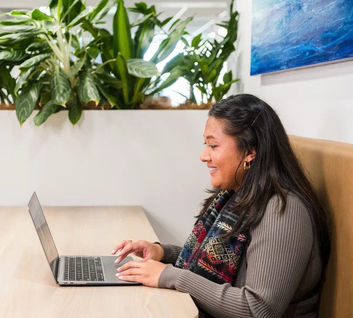 Woman using a laptop, smiling, in a bright office with plants and art.