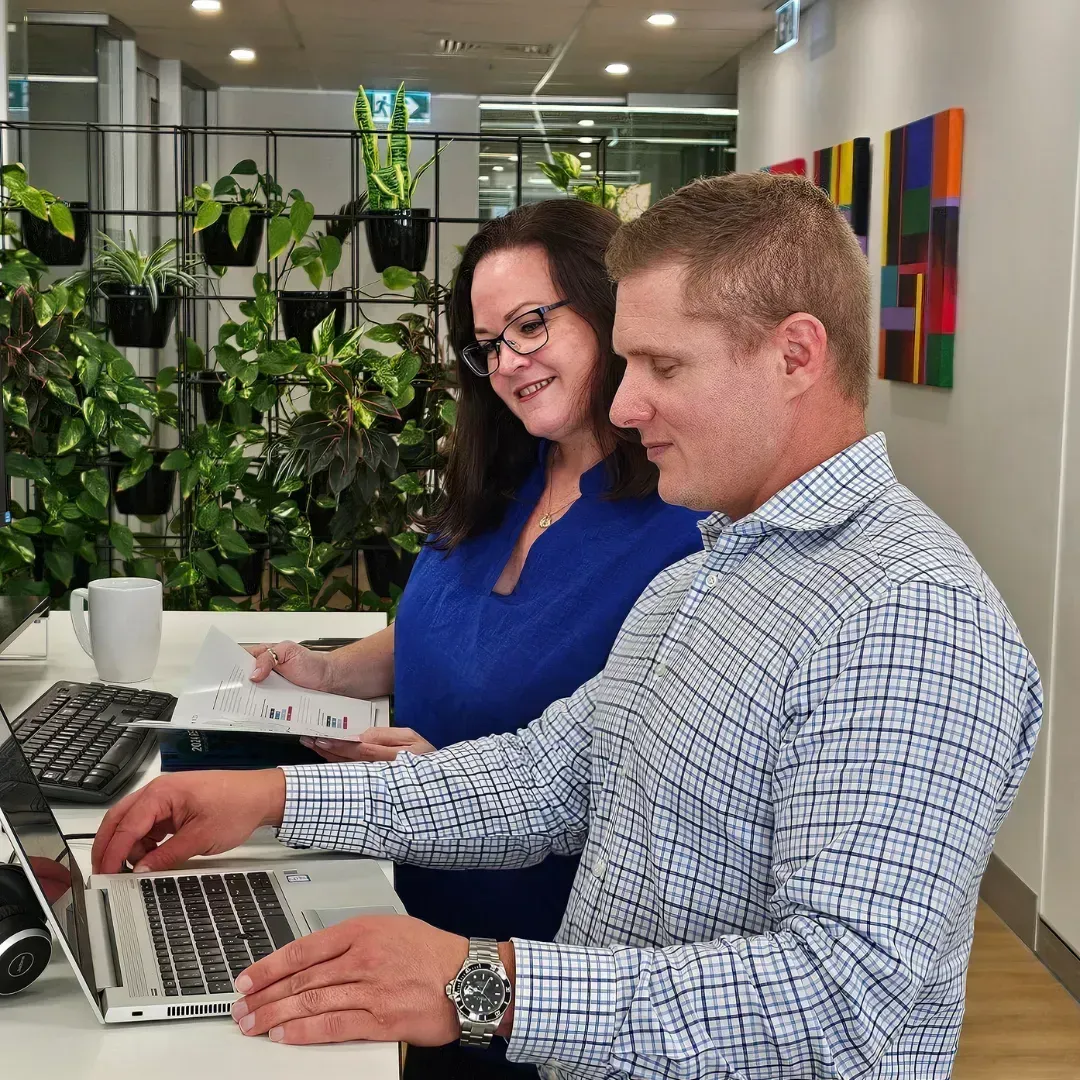 Woman and man looking at a laptop, standing at a desk in an office. Plants and art in the background.