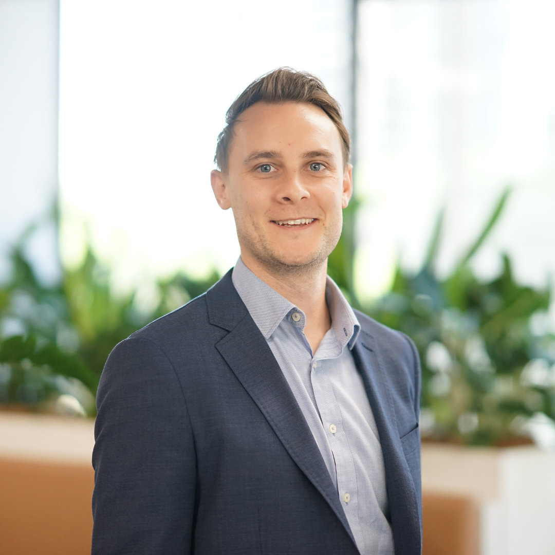 Man smiling in a blue blazer and light blue shirt, standing near plants and a window.