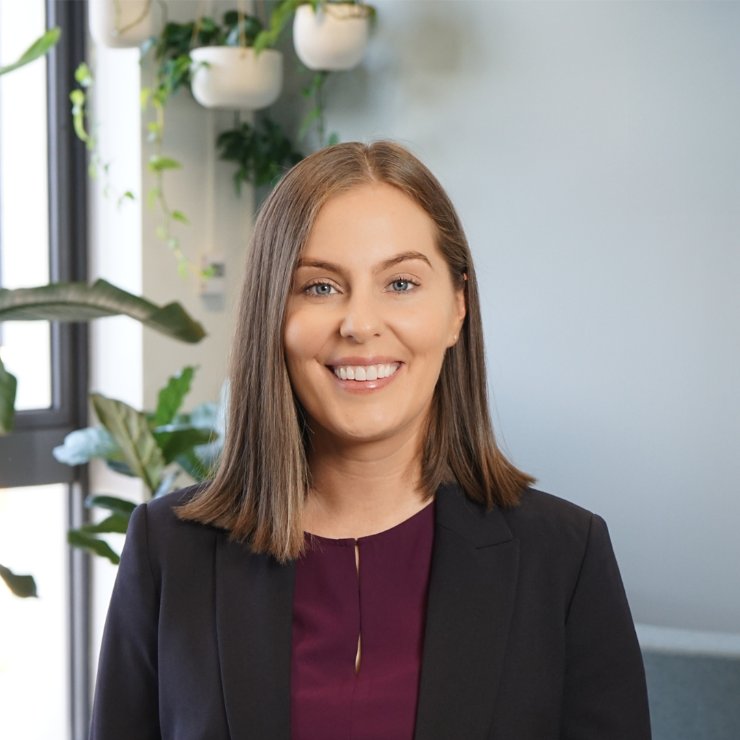 Woman in a dark suit smiles, standing near plants and a window.