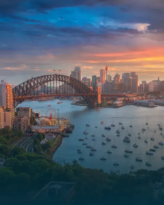 Sydney Harbour at sunset: Harbor Bridge, city skyline, boats, colorful sky.
