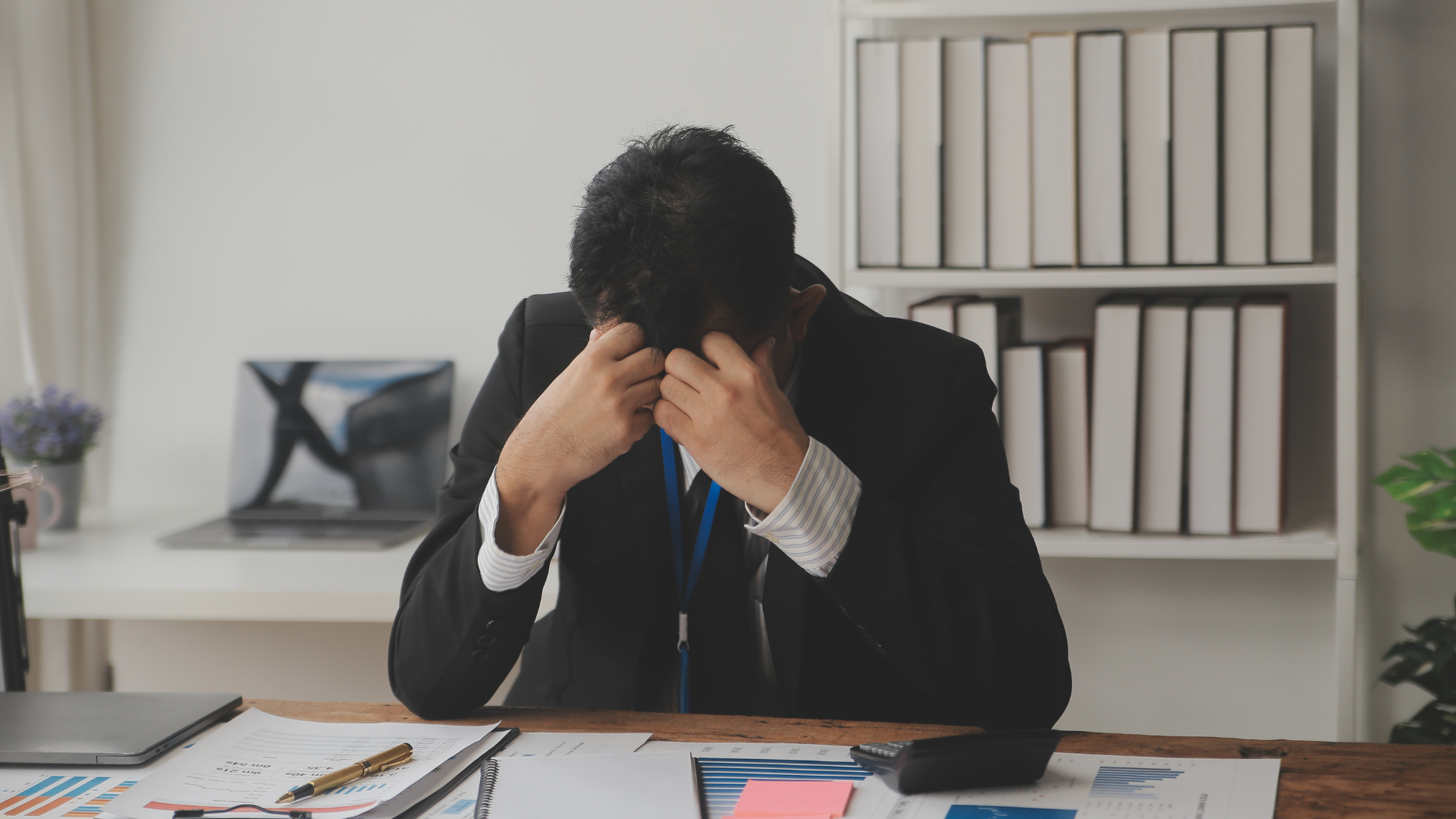 Man in suit stressed, covering face at desk with papers, laptop, and books in a bright office.