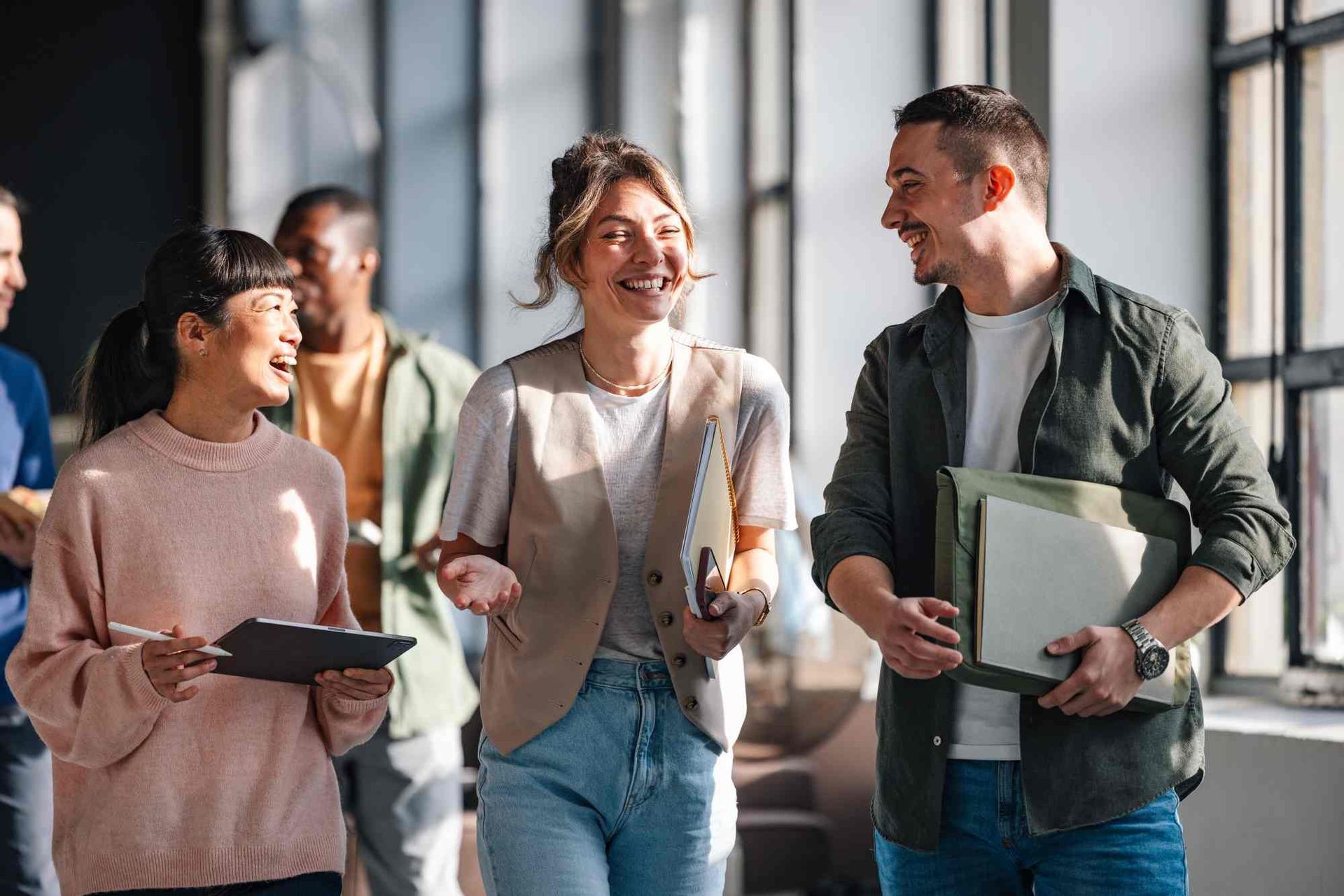 Group of people smiling and talking in a well-lit office hallway, some holding folders and clipboards.