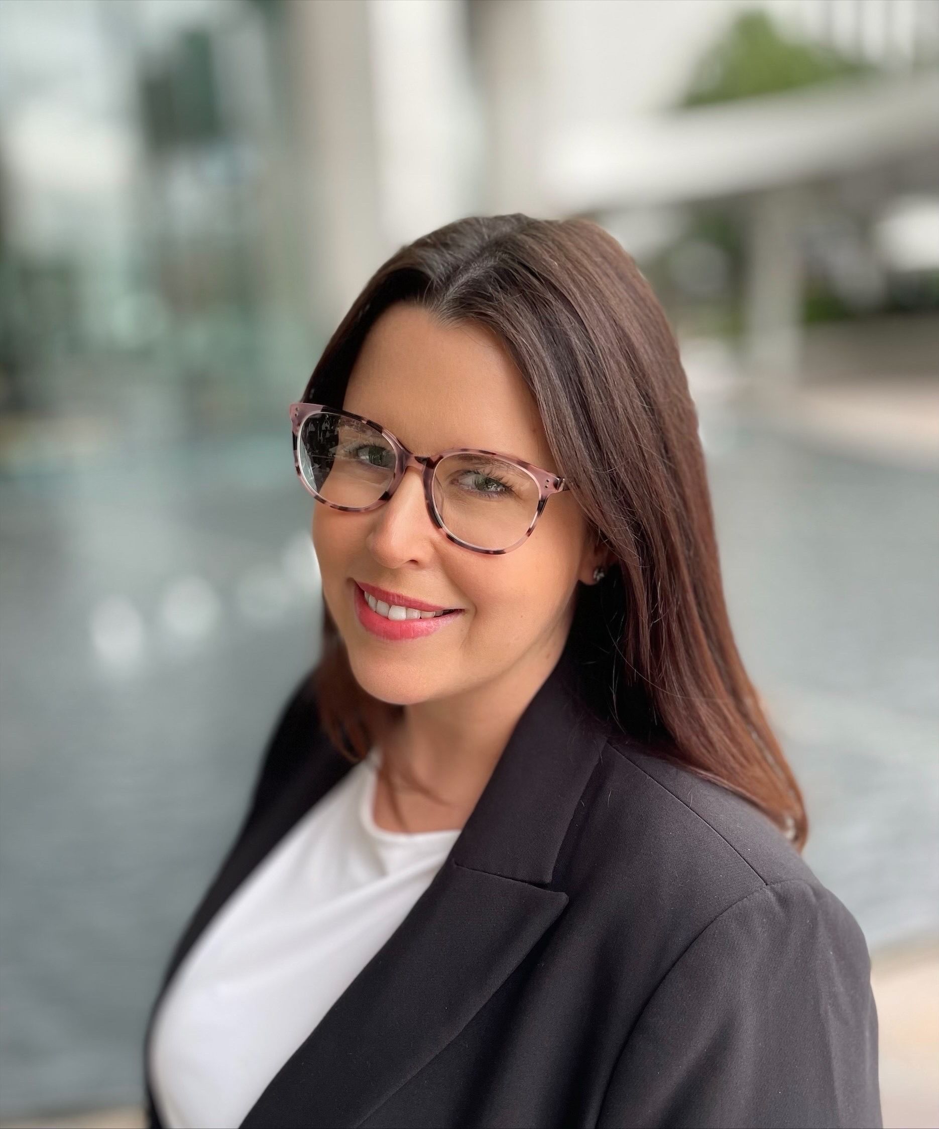 Woman in glasses, black blazer, and white top smiles outdoors near a building.