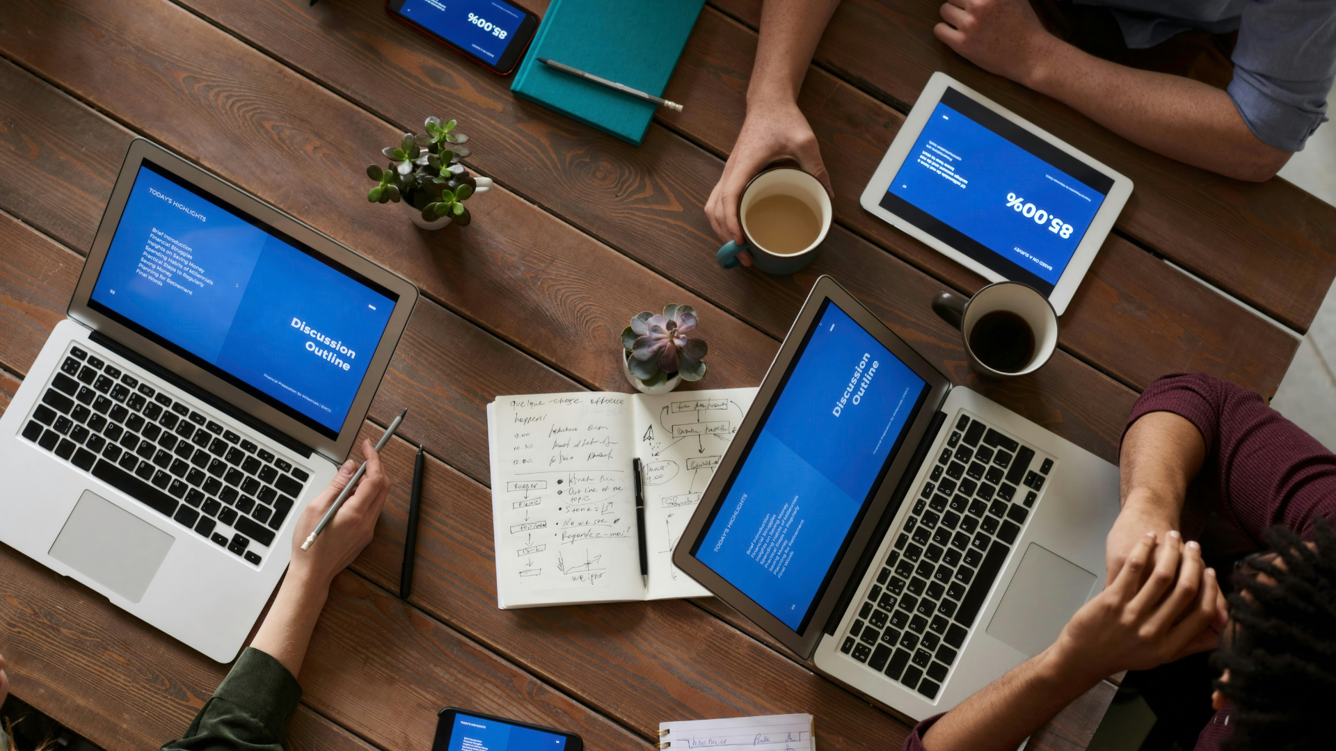 Group of people at a wooden table, laptops and a tablet showing a blue screen. Notebook, plants, and coffee cups.