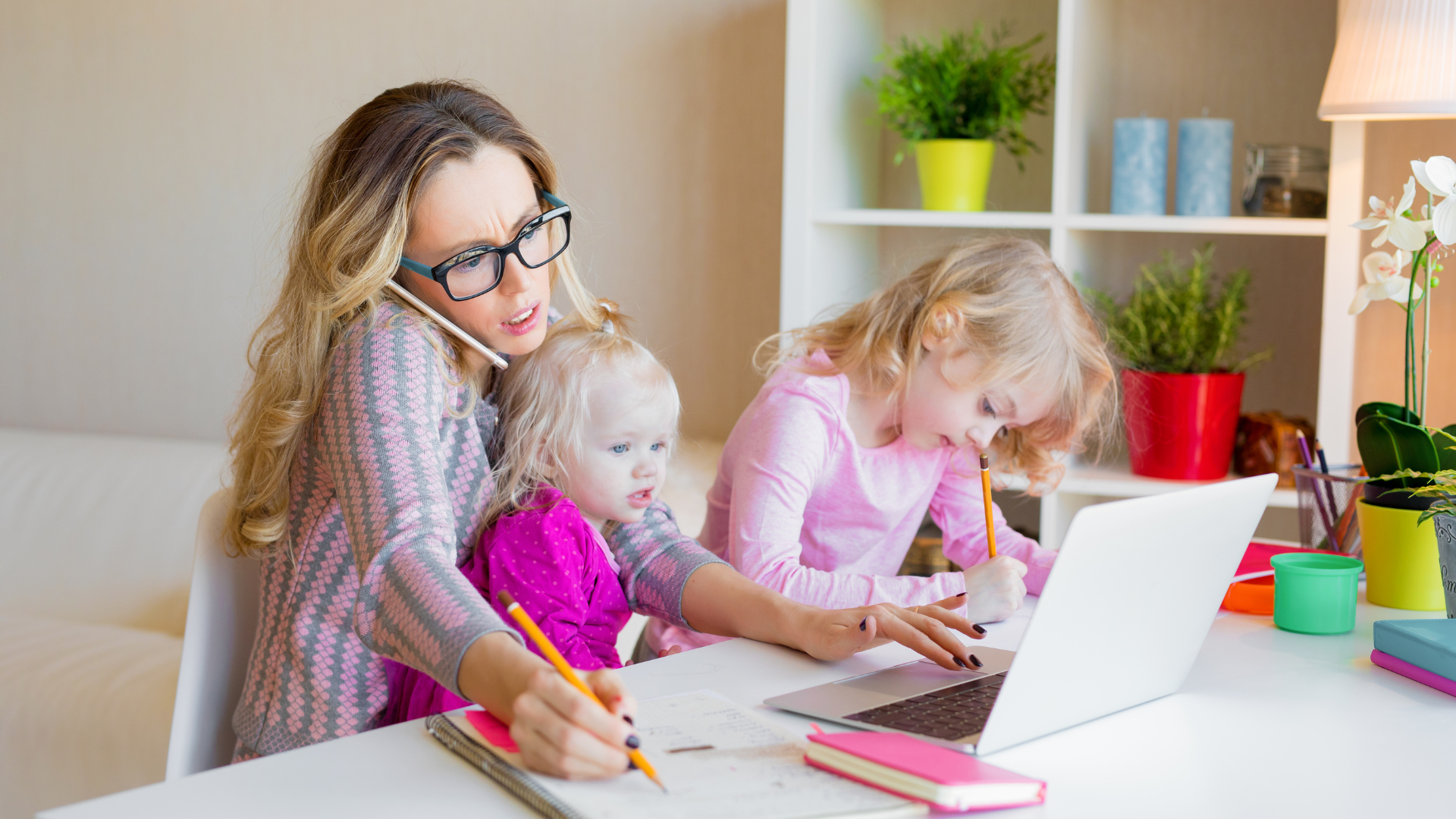 Woman on phone, working on laptop, with two young children. Light-filled room, desk with notebooks, and small plants.