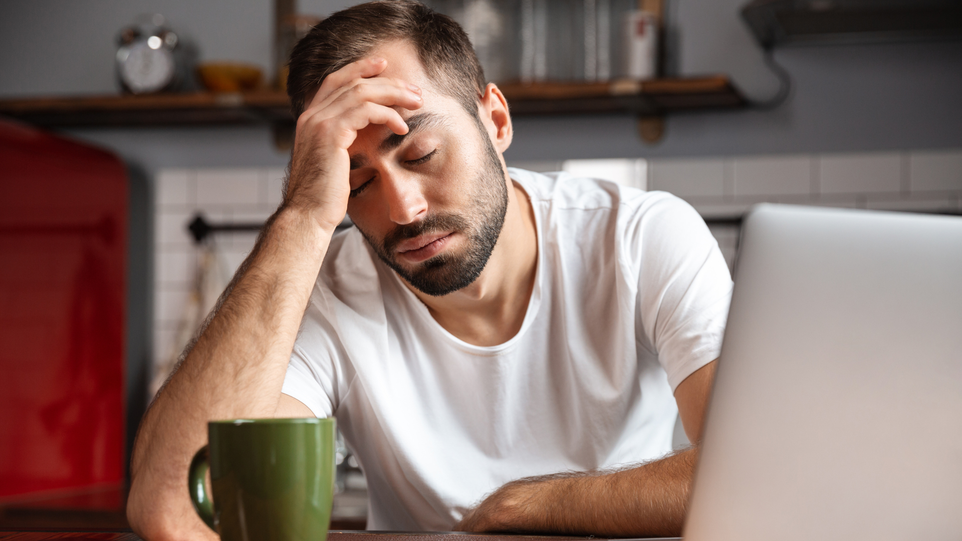 Man with hand on forehead, looking down, coffee mug, laptop, kitchen setting.