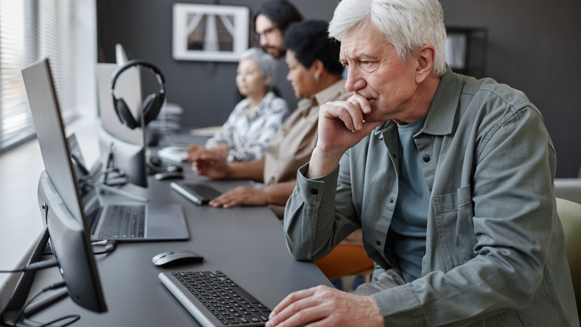 People at computers, focused. Man in foreground touching chin, looking at monitor. Office setting.