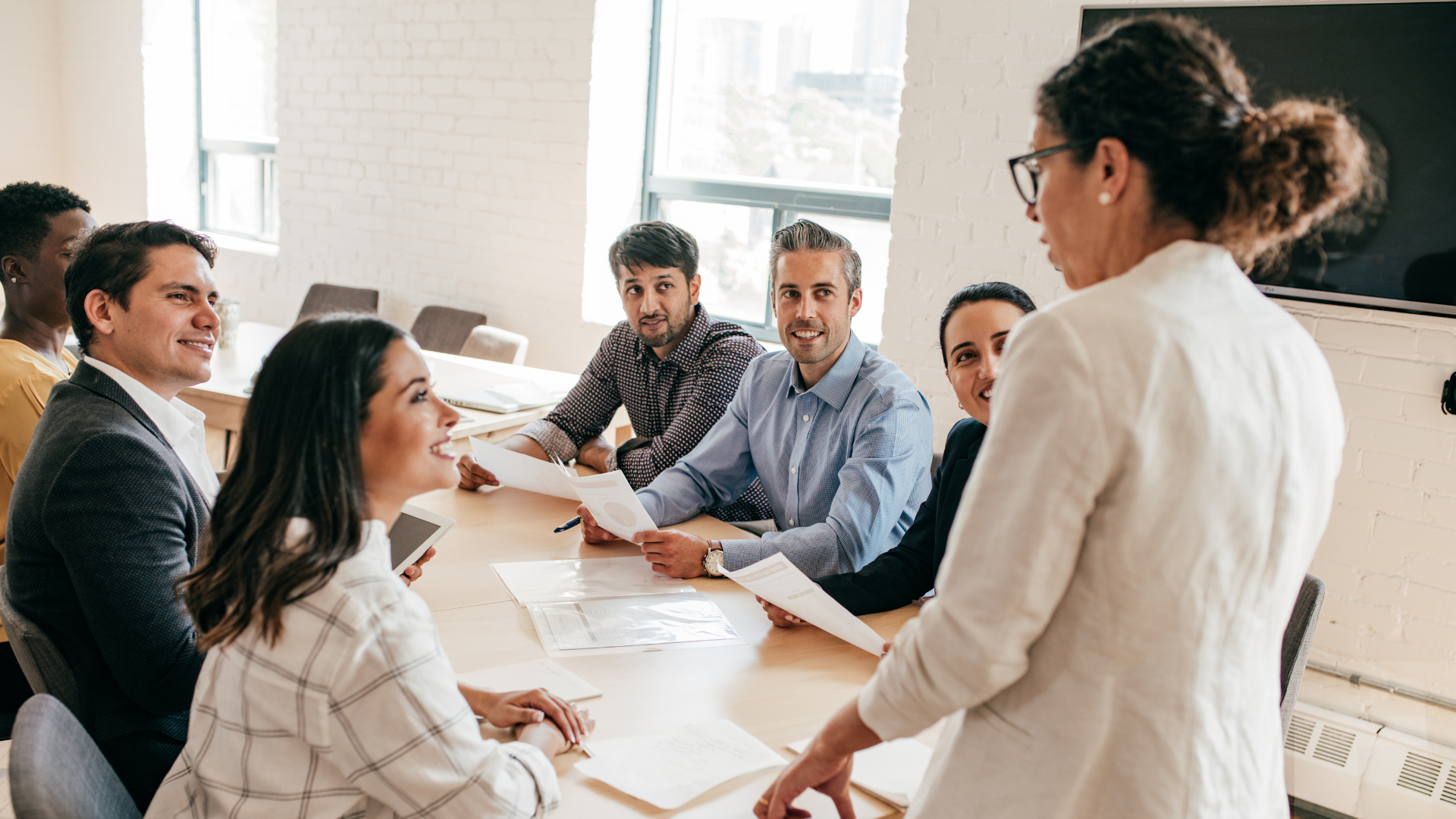 People in a meeting at a conference table, a woman is presenting to the group.