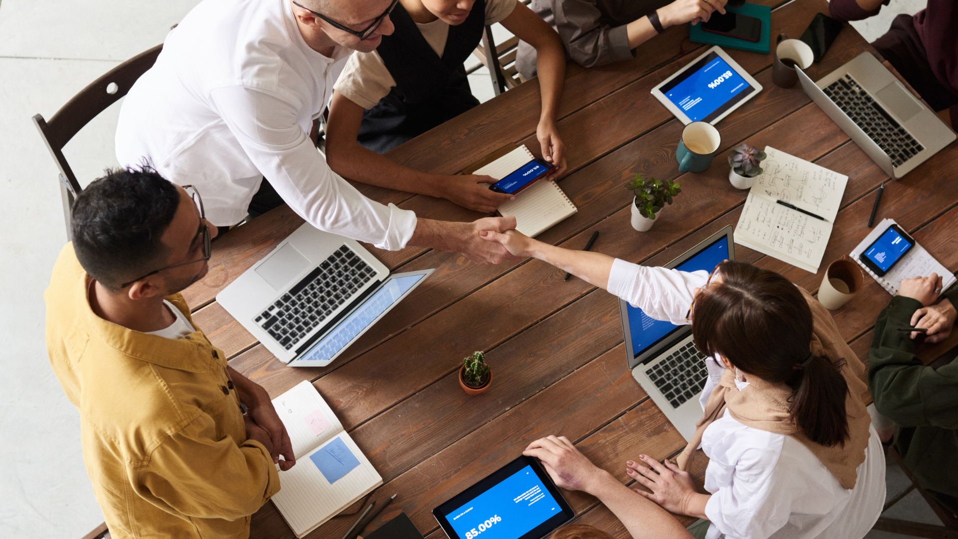 People at a table, two shaking hands, laptops and tablets visible.