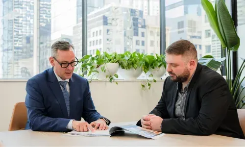 Two men in suits seated at a table, reviewing documents, with a city view in the background.