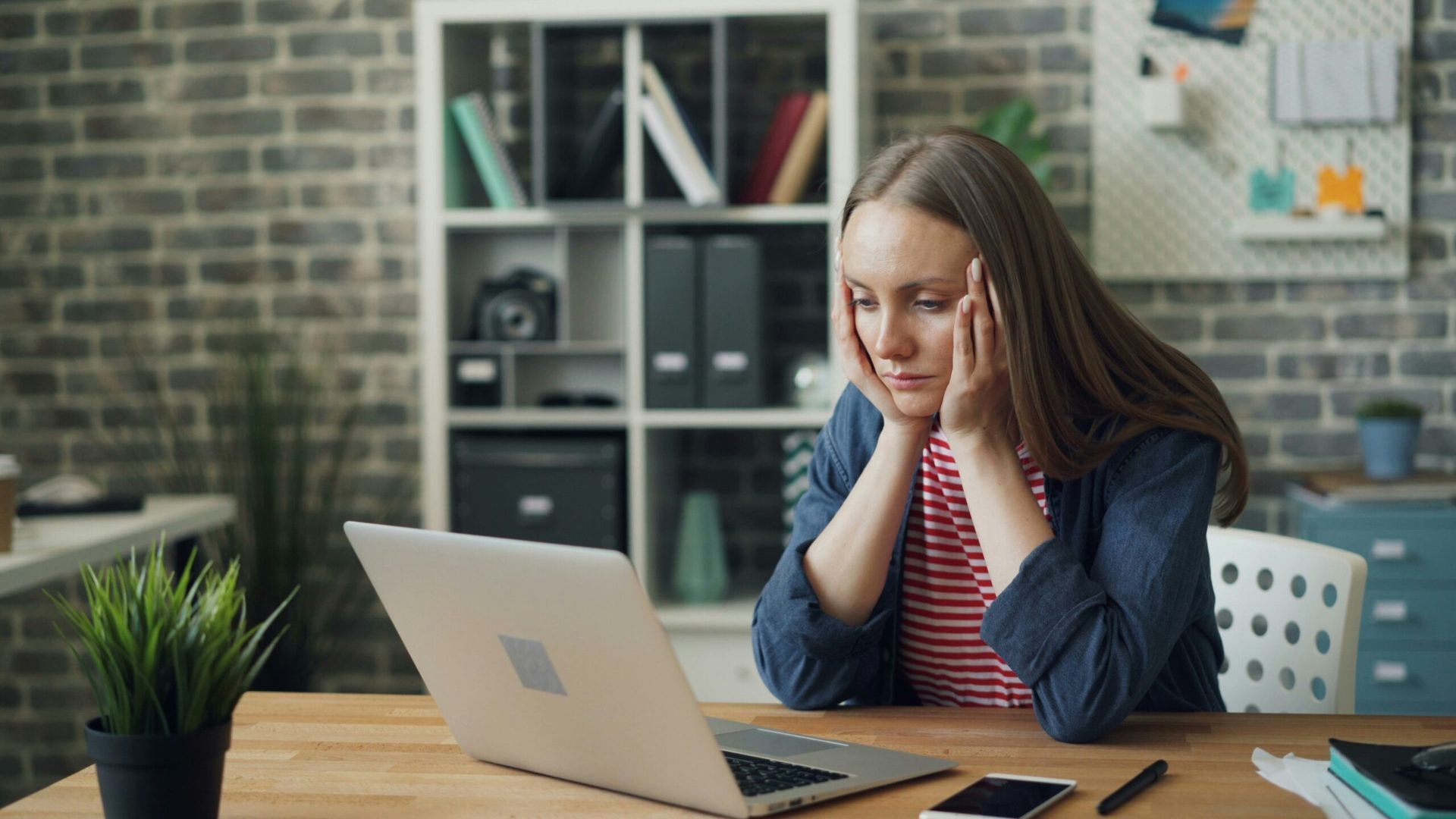 Woman with hands on face, looking at laptop in office, appearing stressed.