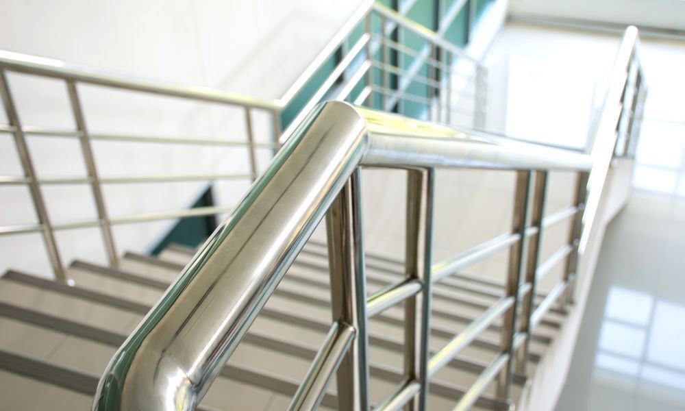 Shiny metal stair railing in a bright indoor stairwell with white steps and glass panels — FBM Fabrication In Dapto, NSW