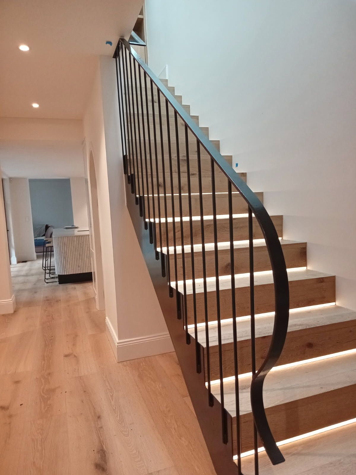 Modern staircase with black railing and illuminated wooden steps in a bright hallway  — FBM Fabrication In Dapto, NSW