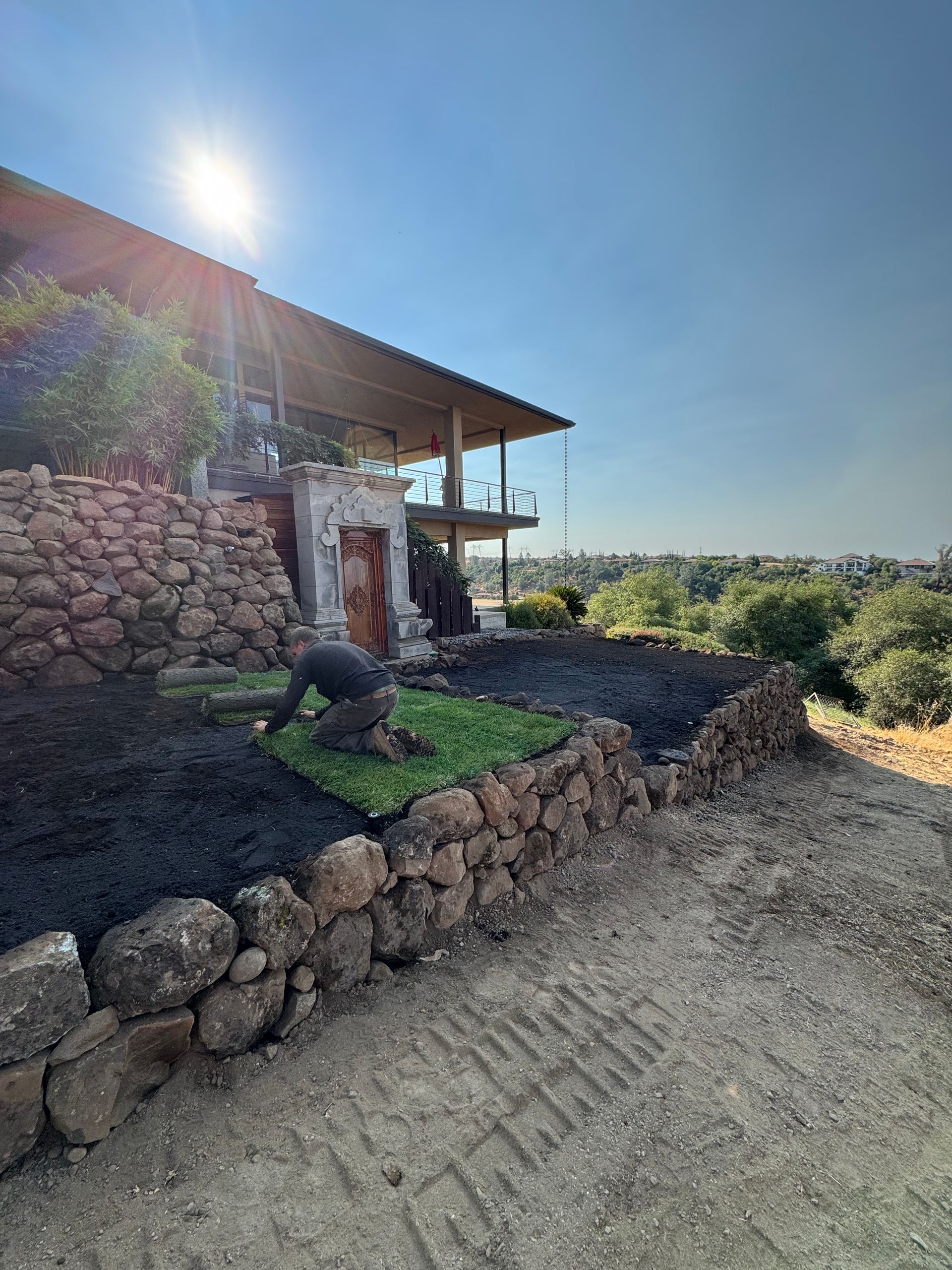 Person laying sod near a stone wall and a modern house under a bright sun.