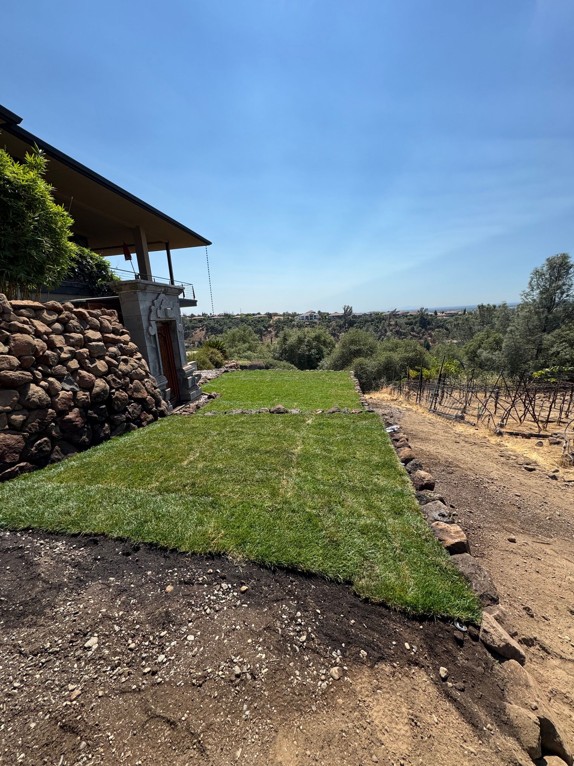 Freshly laid green sod beside a house with a stone wall, overlooking a sunny landscape.