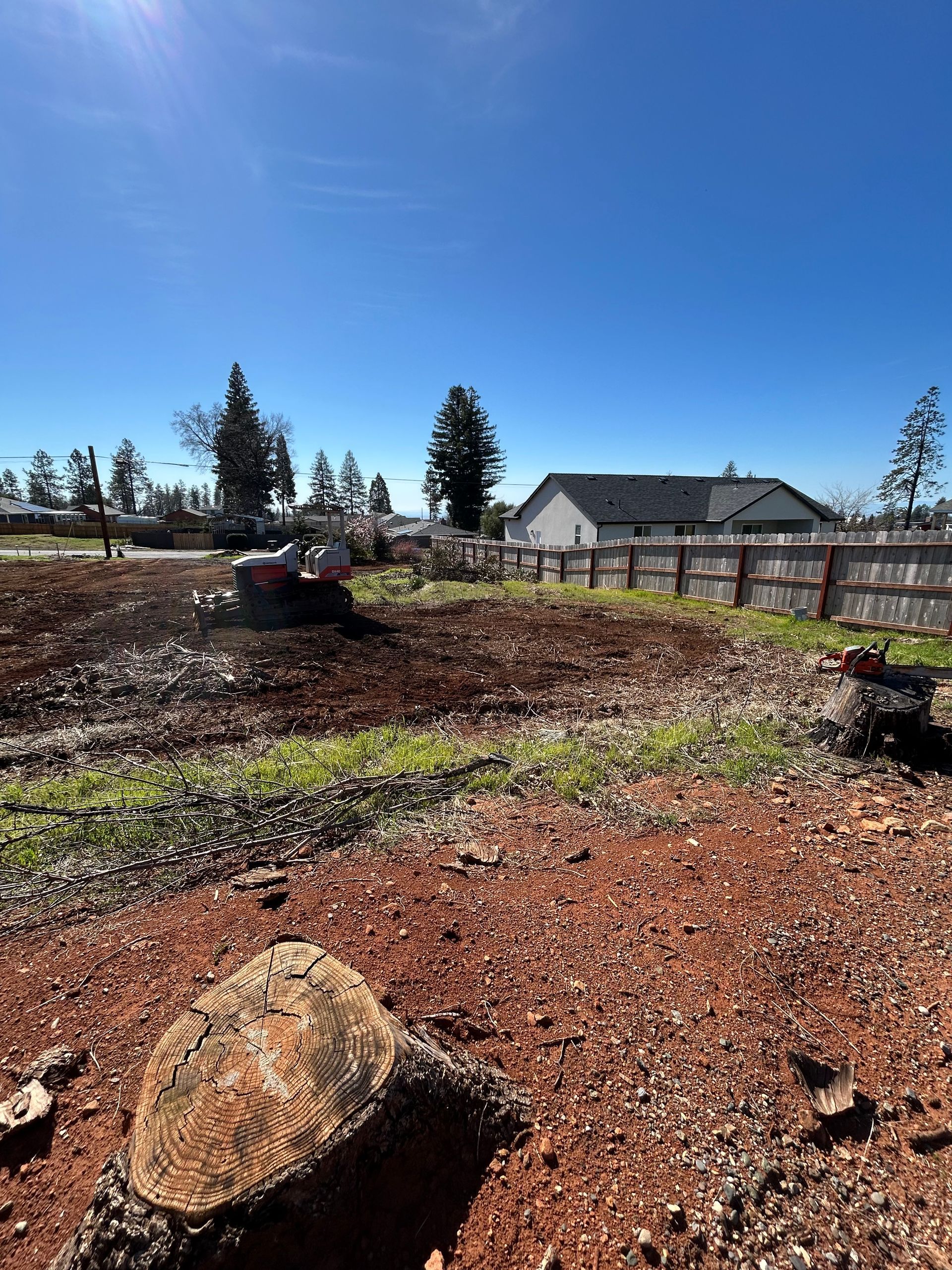 Brown land being cleared with equipment on a sunny day, a house and trees in the background.