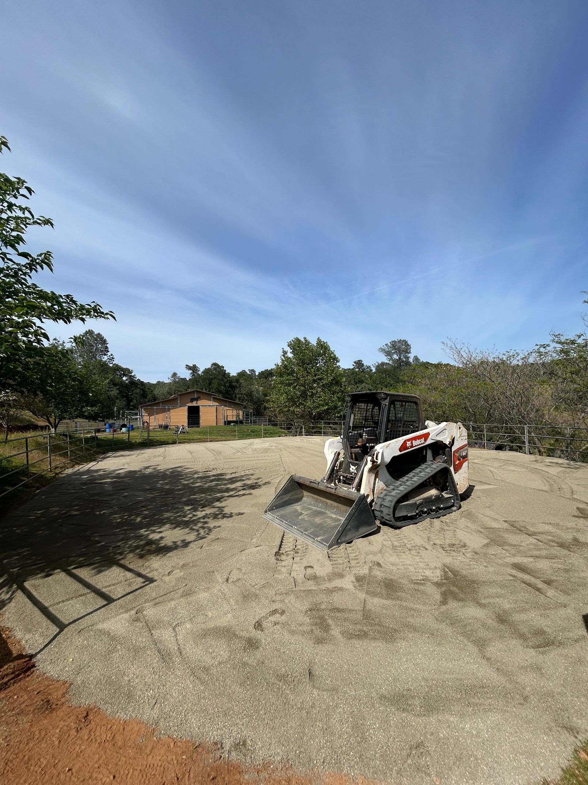 Skid steer on gravel, clearing an area near a barn and trees under a partly cloudy sky.