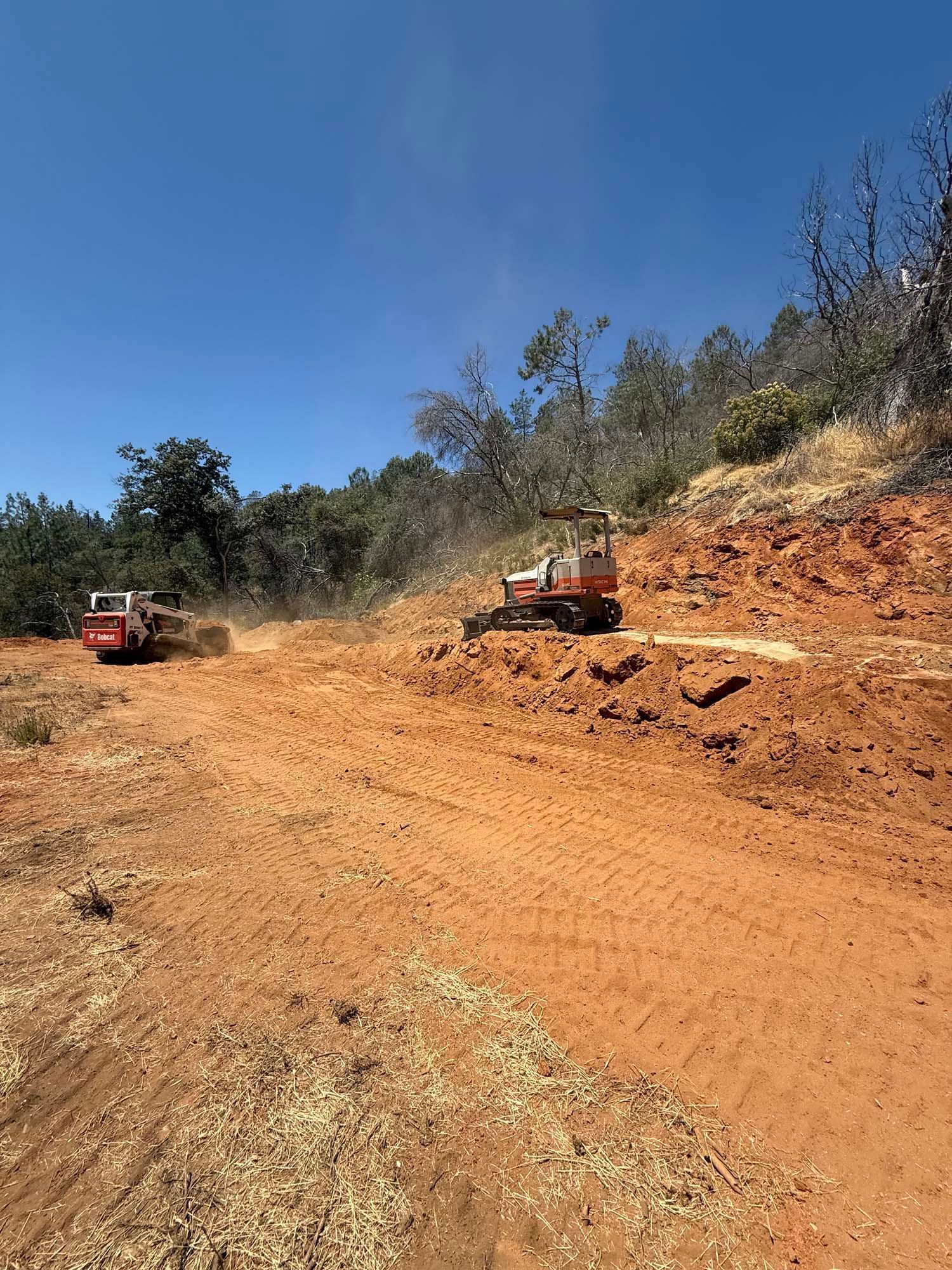Two small orange construction vehicles on a dirt hillside, under a bright blue sky.