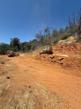 Two orange machines clearing a dirt hillside, kicking up dust under a blue sky.